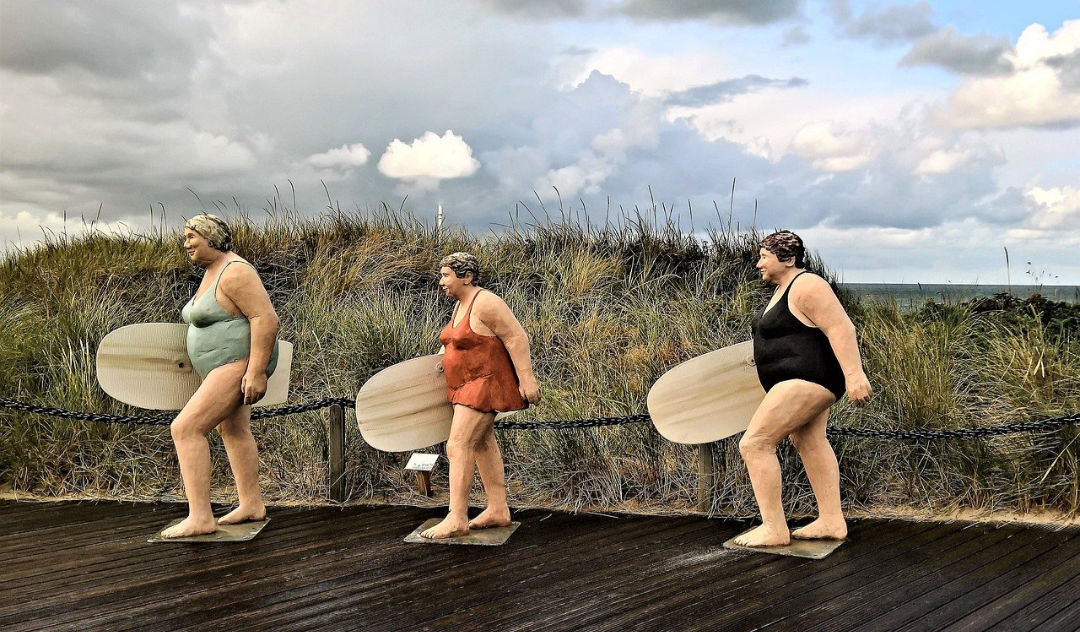 Three old ladies in swimsuits carrying paddle boards on a boardwalk next to a grass swept and dune covered beach. Sculpture. Women of a certain age.