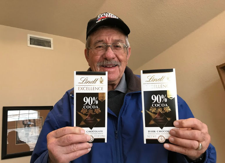 Senior man in baseball cap holding up two Lindt 90% chocolate bars with a big grin. The power of small gestures.
